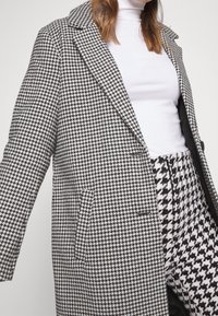 Abrigo de patrón de espiga en blanco y negro con cuello con muescas, diseño de botón simple y dos bolsillos frontales. Textura suave con un ajuste estructurado.