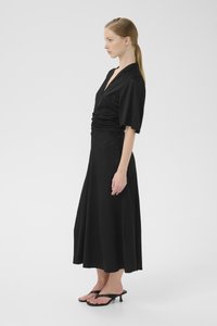 Black dress with a wrap design, short sleeves, ruched waist, flowing skirt, and textured fabric. Paired with black heeled sandals.