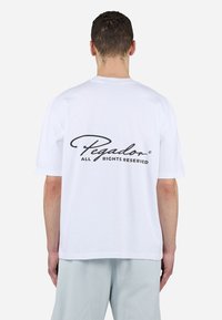Camiseta de algodón blanca con un logo en cursiva negro en la parte superior de la espalda, mangas cortas y un corte relajado. El texto dice "Pegador Todos los Derechos Reservados."