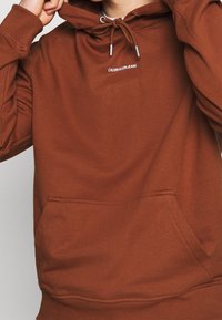 Felpa marrone in pile con tasca a marsupio, cappuccio con coulisse e logo bianco "CALVIN KLEIN JEANS" sul petto. Texture morbida.