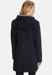 Navy wool coat featuring a hood, tailored design, and back vent. Textured fabric with a clean silhouette and subtle seams.