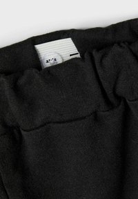 Schwarzer Stoffbund mit elastischen Rüschungen, versehen mit einem weißen Etikett, auf dem ein Logo und ein kleines kreisförmiges Akzent zu sehen sind. Weiche Textur.