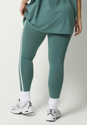 Persona con leggings color verde petrolio con strisce laterali bianche, calzini bianchi e scarpe da ginnastica New Balance bianche, in piedi con una mano sul fianco.