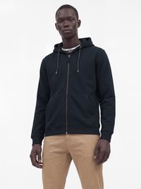 Homme portant un sweat à capuche noir zippé avec des cordons et un pantalon beige, debout devant un fond blanc uni.