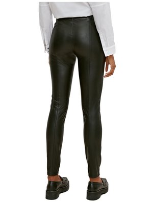 Takakuvassa henkilö, jolla on mustat tiukat nahkahousut, valkoinen pitkähihainen paita ja mustat paksupohjaiset loafersit, seisomassa valkoisella taustalla.