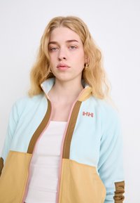 Jachetă cu fermoar de culoare bleu deschis și bej, cu accente și logo roz, având o textură moale și un design în blocuri de culoare.
