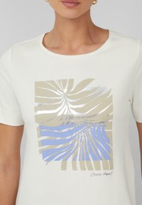 T-shirt à manches courtes, couleur crème, avec un design graphique mettant en valeur des motifs de feuilles beiges et bleus ainsi que des accents argentés. Encolure ronde.