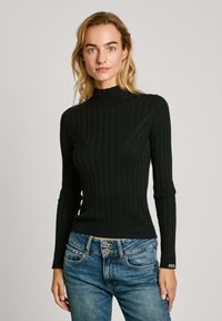 Svart ribbad polotröja med långa ärmar och en figurnära design, parat med blå denimjeans som har guldknappdetaljer.