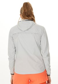 Lys grå, letvægtsjakke med hætte, der har en mesh-rygpanel og lange ærmer, båret over klare orange shorts.