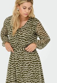 Vrouw die de tailleband aanpast van een lange mouw olijfgroene en beige gestreepte jurk met volants aan de mouwen, tegen een effen lichte achtergrond.