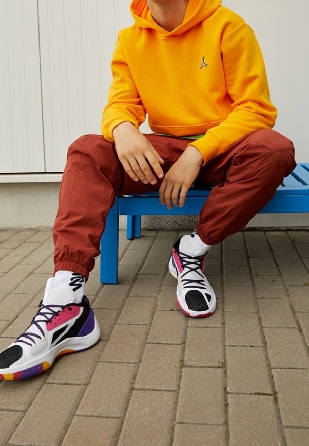 Sportovní boty s černou a bílou síťovinou, s akcenty v růžové, fialové a oranžové barvě. Model nosí oranžovou mikinu a červené tepláky.