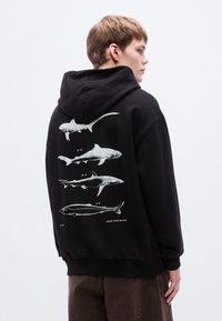 Μαύρο φούτερ με κουκούλα που έχει λευκές απεικονίσεις πέντε ειδών καρχαριών στην πλάτη, με λεπτομερείς ετικέτες. Υφασμάτινο από βαμβάκι, με άνετη εφαρμογή.