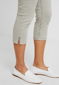 Pantaloni grigio chiaro a cropped con orlo risvoltato e dettaglio a bottone, abbinati a scarpe bianche senza lacci con una texture in pelle liscia.