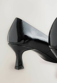 Scarpa nera lucida con tacco alto angolato per mostrare il tacco affusolato e la punta appuntita, su uno sfondo neutro.