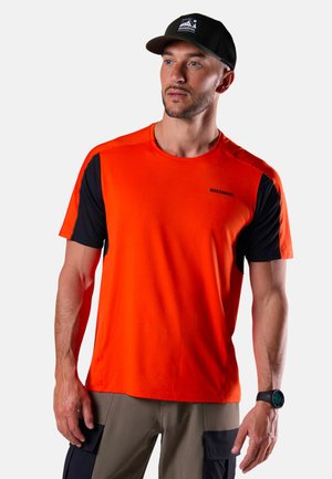 Uomo adulto che indossa un berretto nero, una maglietta a maniche corte arancione brillante e nera, pantaloni beige con tasche nere e uno smartwatch nero, in piedi fermo.