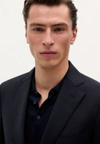 Blazer azul marino sobre una camiseta polo oscura, con un diseño estructurado, tela suave y detalles de costura sutiles. Silueta limpia y sencilla.