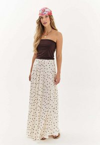 Žena oblečená v tmavohnedom topu bez ramienok, s dlhou bielou sukňou s čiernymi bodkami, kvetinovým šatkou na hlave a sandálmi, stojí pred bielym pozadím.