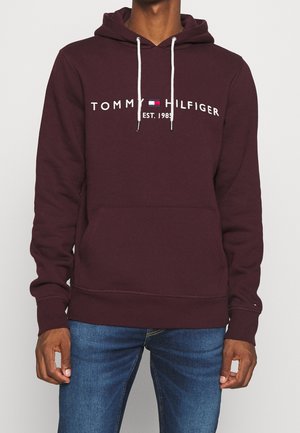 Man draagt een bordeauxrode Tommy Hilfiger hoodie met witte trekkoorden en logo, gecombineerd met een blauwe spijkerbroek, staand tegen een effen achtergrond.