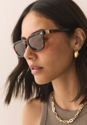 Középhosszú sötét hajú nő, aki teknősbéka mintás napszemüveget, arany láncot és arany csepp alakú fülbevalókat visel.