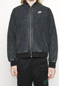 Svart denim bomberjacka med dragkedja framtill, ribbade muddar och krage, med en vit Nike-logotyp på bröstet. Avslappnad passform.