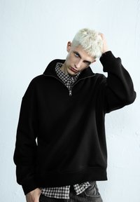 Svart zip-up sweatshirt med hög krage, ribbade muddar och texturerat tyg, lager-på-lager över en rutmönstrad skjorta, bärs med mörka jeans.