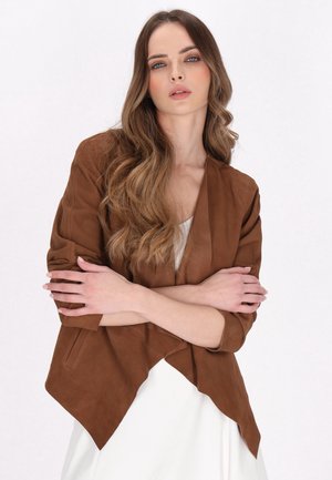 Junge Frau mit langen welligen Haaren, die eine braune Jacke und ein weißes Kleid trägt, steht mit verschränkten Armen vor einem weißen Hintergrund.