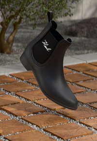 Mustad mustad Chelsea saapad, valmistatud kummist, millel on matta viimistlus, elastsed külgpaneelid ja valge logo välisküljel, kuvatud õhus.