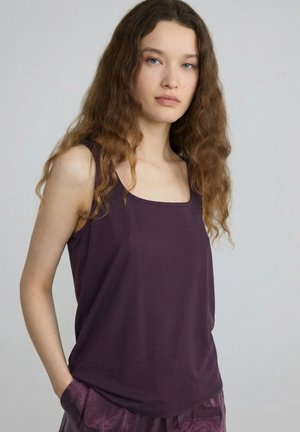 Jonge vrouw met lang golvend haar, gekleed in een mouwloze donkerpaarse top en bijpassende broek, staand tegen een effen lichte achtergrond.