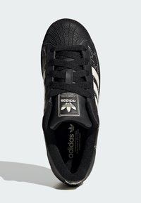 Black sneaker with a textured rubber toe cap, black fabric upper, white stripes, and a black logo patch. Laces are black and textured.