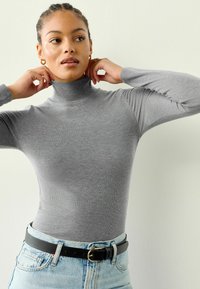 Grauer Rollkragenpullover aus geripptem Stoff, taillierte Passform, lange Ärmel, getragen mit einem schwarzen Gürtel über hellblauen Jeans.