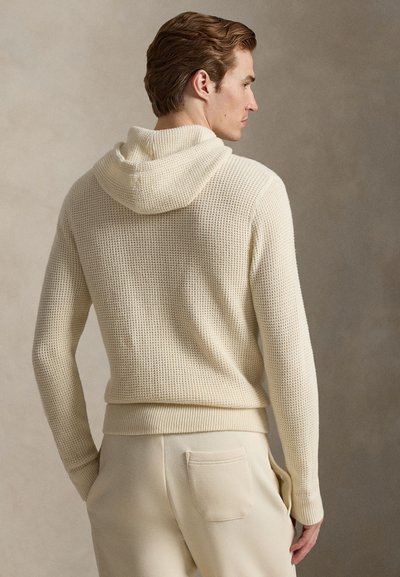 Vaaleanbeige neulottu huppari, jossa on teksturoitu kudos, tilava huppu sekä resorit hihansuissa ja helmassa. Yhdistetty vaaleanbeigeihin housuihin, joissa on takatasku.