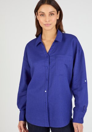 Femme portant une chemise bleu roi ample, à manches longues, boutonnée, avec une poche poitrine, debout devant un fond clair uni.