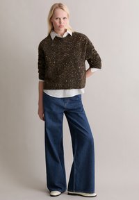 Brązowy sweter z wzorem, założony na białą koszulę zapinaną na guziki, zestawiony z szerokimi dżinsami w kolorze niebieskim i białymi botkami do kostki. Jednoliste tło.