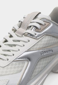 Λευκό και ασημί αθλητικό παπούτσι Calvin Klein με ύφασμα από δίχτυ, συνθετικές επενδύσεις, λευκά κορδόνια και κεντημένη ετικέτα στο γλωσσίδιο.