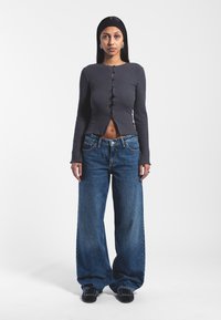 Szary prążkowany krótki top z guzikami i długimi rękawami, z rozcięciem z przodu, połączony z luźnymi, niebieskimi dżinsami o wysokim stanie. Czarne buty.