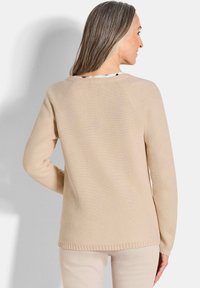 Frau mit langen grauen Haaren, die einen beigen, strukturierten Strickpullover und helle Hosen trägt, steht vor einem schlichten weißen Hintergrund und schaut abgewandt.