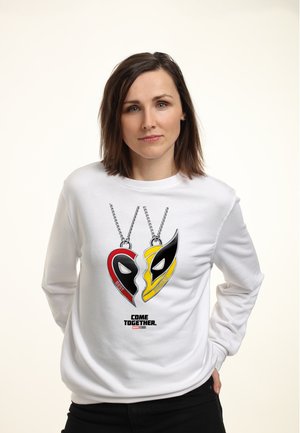 Kvinde iført en hvid sweatshirt med et design af et rødt og gult brudt hjerte-halskæde mærket "Bedste Venner" og teksten "Come Together Marvel Studios".