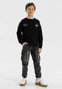 Zwarte sweatshirt met een witte halslijn, voorzien van een blauw en rood logo. Grijze cargobroek met meerdere zakken. Witte sneakers met rode accenten.