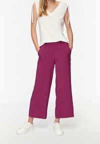 Femme portant un haut sans manches blanc, un pantalon large magenta et des baskets blanches, se tenant les mains dans les poches contre un fond uni.