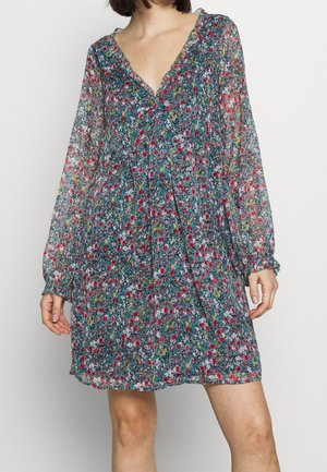 Vrouw die een bloemenjurk met lange mouwen draagt die boven de knie valt, met een V-hals en geplooide details bij de borst, staand tegen een effen achtergrond.