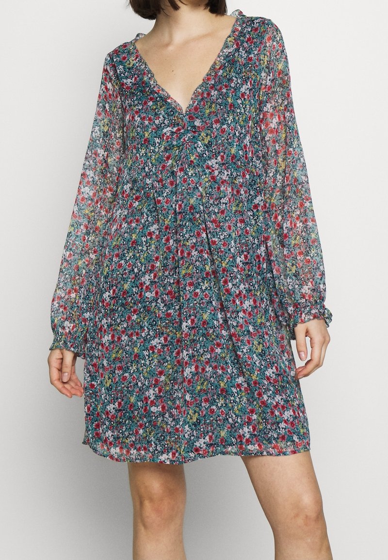 Femme portant une robe fleurie à manches longues, au-dessus du genou, avec un col en V et un détail froncé au niveau de la poitrine, debout devant un fond uni.