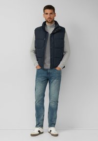 Granatowa kamizelka puchowa z wysokim kołnierzem, na jasnoszarym swetrze z golfem. Do tego wyblakłe niebieskie dżinsy i białe trampki z szarymi akcentami.