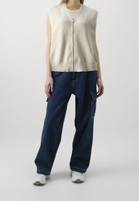 Hellbeige ärmellose Zip-Weste mit gerippter Textur, kombiniert mit locker sitzenden dunkelblauen Cargo-Jeans. Weiße Sneakers runden das Outfit ab.