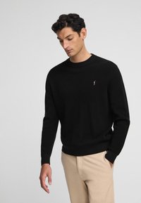 Suéter negro de punto con cuello redondo, puños y dobladillo acanalados, con un pequeño y sutil logotipo de la marca en el pecho. Lleva puesto un pantalón beige.