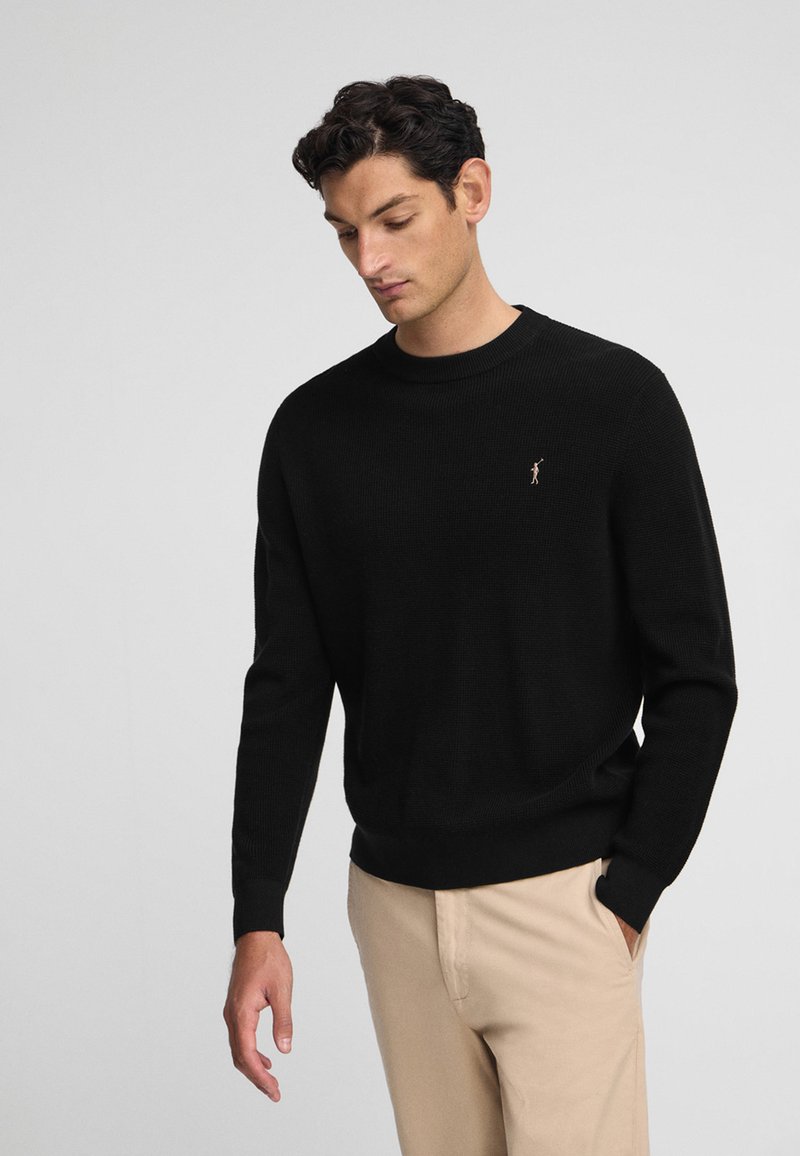 Suéter negro de punto con cuello redondo, puños y dobladillo acanalados, con un pequeño y sutil logotipo de la marca en el pecho. Lleva puesto un pantalón beige.