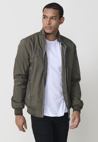 Olīvu zaļš bomber žakets ar rāvējslēdzēju priekšā, ribotiem piedurknēm un sānu kabatām; uz krūtīm ir neliels logotips un gluda tekstūra.