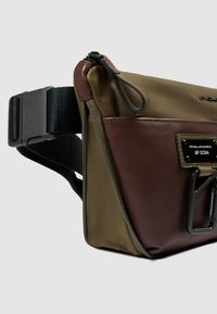 Bolso bicolor que combina cuero verde oliva y marrón, diseño rectangular, superficie texturizada y una correa negra ajustable con hebilla de plástico.