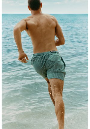 En mand i grønne, stribede badeshorts løber ud i havet med vand, der sprøjter omkring hans ben under en overskyet himmel.