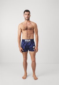 Jūrų jūrų satino šortai su juodu elastingu juosmeniu, kuriuose priekyje yra baltas Venum logotipas. Modelis stovi basomis, užimančiu neutralią pozą.