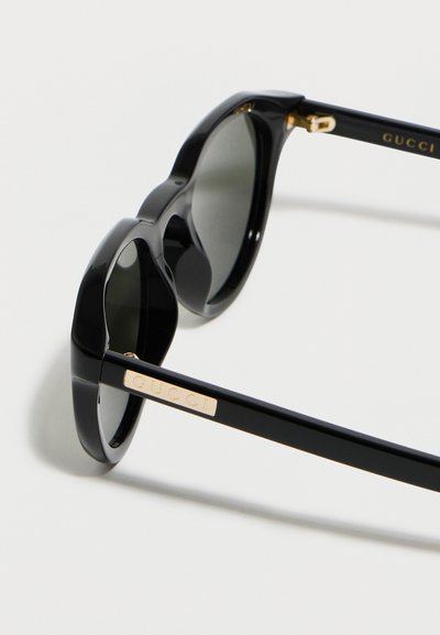 Γυαλιά ηλίου σε σχήμα γάτας μαύρου χρώματος με σκουρόχρωμους φακούς και χρυσό λογότυπο Gucci στα πλαινά, εμφανίζονται σε γωνία πάνω σε λευκή επιφάνεια.